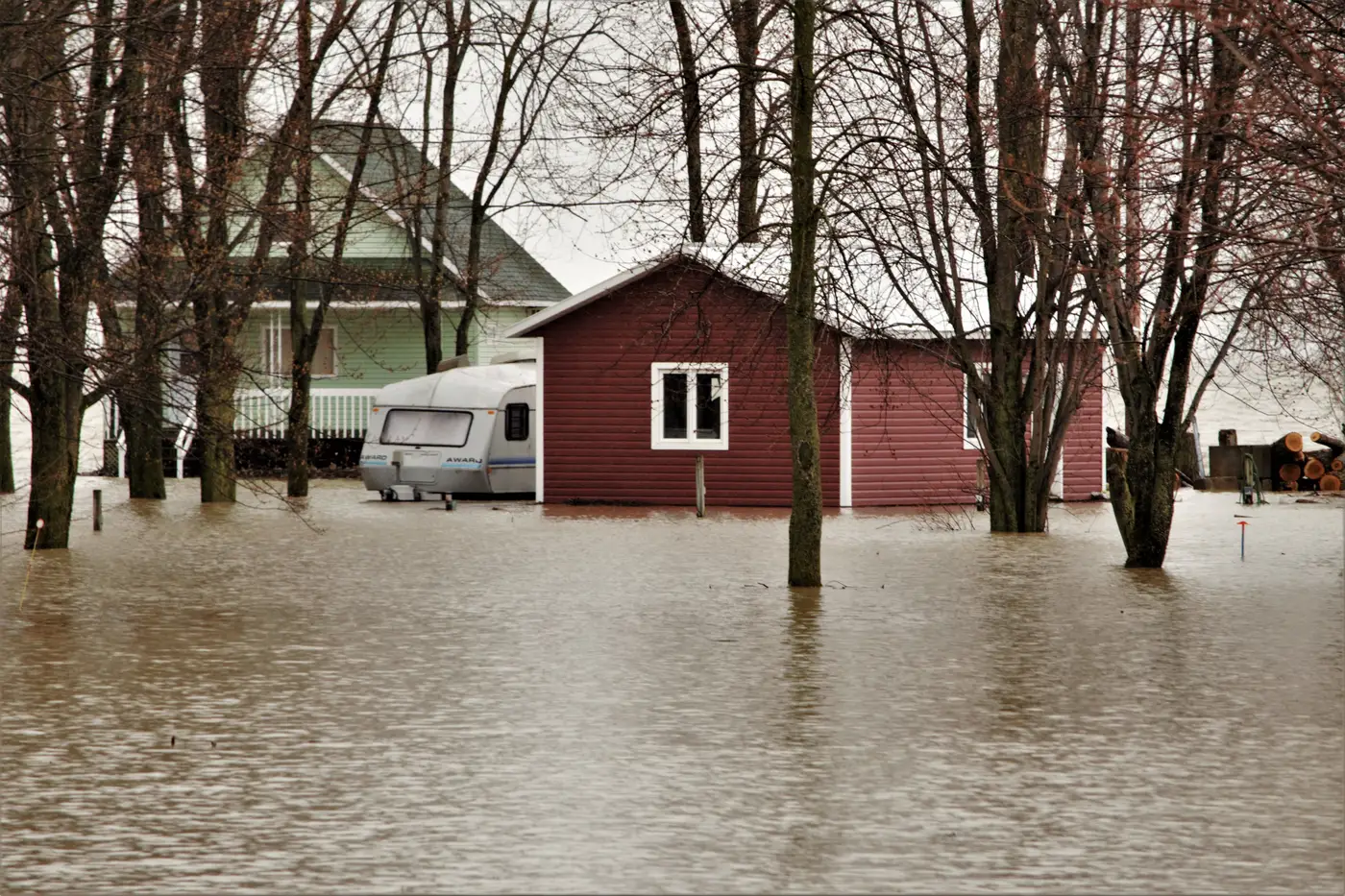 A red cabin and a small camper trailer surrounded by floodwater among bare, leafless trees.