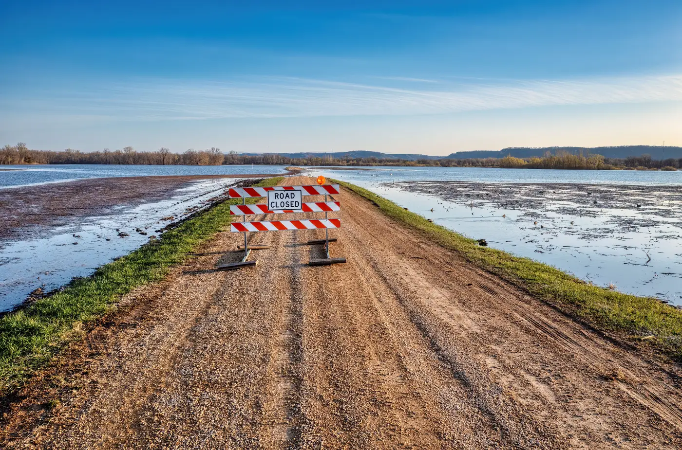 A ROAD CLOSED sign on a gravel road beside a calm, wide flooded field under a clear sky.